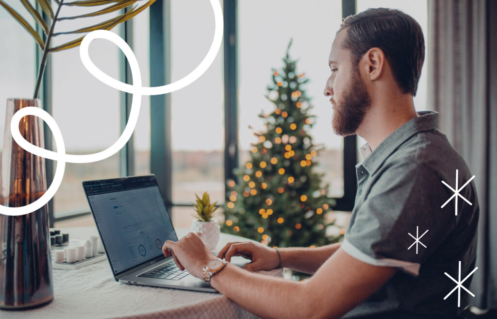 Person working on a laptop at a table near a decorated Christmas tree, with abstract white line and star graphics overlayed.