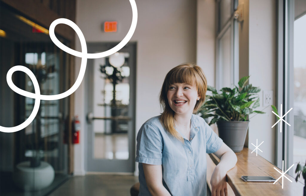 A smiling woman with short light-brown hair sits at a wooden counter by a window, looking to her left. A potted plant and a smartphone rest beside her. The space appears to be a modern office or café with soft natural light, a glass door in the background, and an exit sign visible. White decorative doodles (curved lines and star shapes) are overlaid on the image.