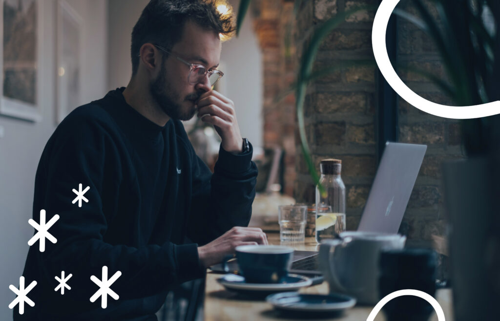 Man with glasses working thoughtfully on a laptop at a café table with coffee and water nearby.