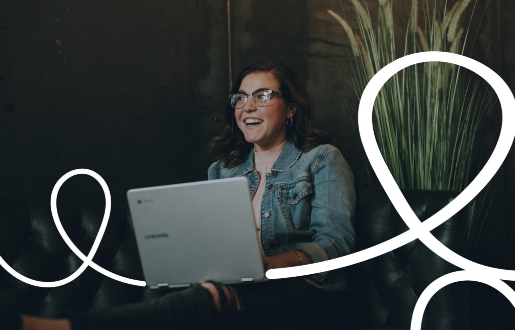 A woman wearing glasses and a denim jacket smiles while working on a laptop, seated on a dark couch with a tall plant in the background. White looping graphic lines overlay the image.