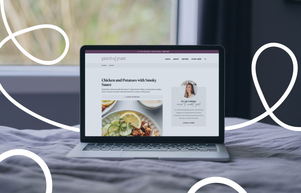 A laptop resting on a bed displays a food blog webpage titled “Chicken and Potatoes with Smoky Sauce” from “Pinch of Yum.” The screen shows a recipe with an image of the dish and a sidebar featuring the site’s author. The background is softly blurred, with a window behind the laptop, and decorative white line doodles overlay the image.